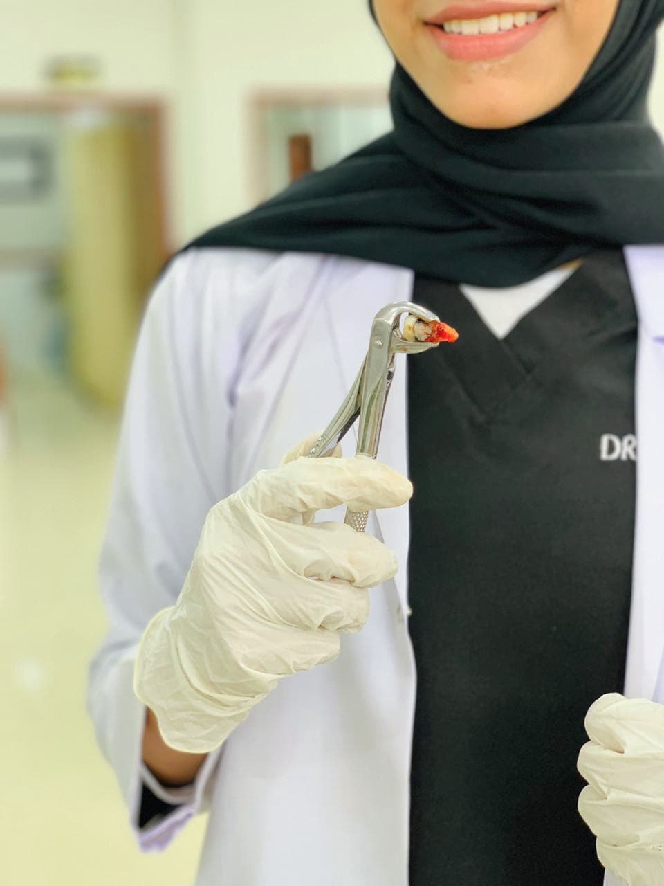 Smiling dentist wearing a hijab holds a bloody extracted tooth with metal dental forceps.