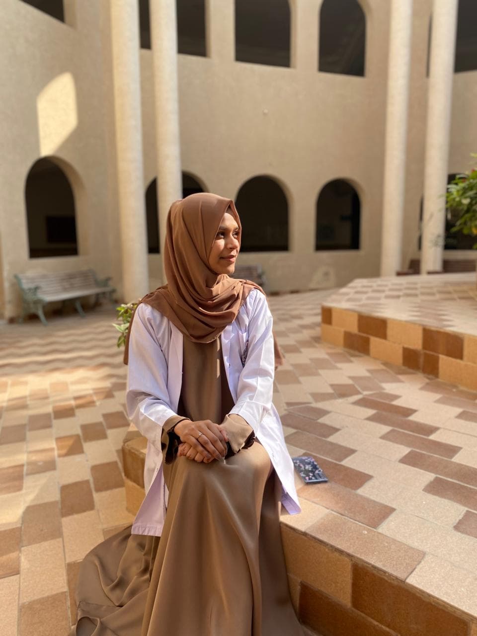 Woman wearing a brown hijab and white lab coat sits in a sunlit courtyard.